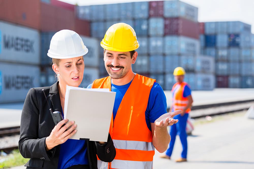 Manager with clipboard full of freight documents talking with worker on shipment yard in front of container